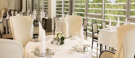 A dining area with round tables set for a meal, featuring white tablecloths, silverware, and candles. Large windows provide a view of greenery outside.