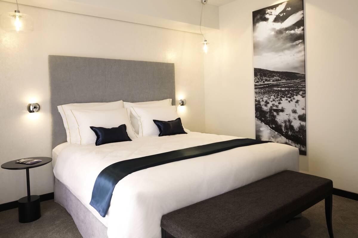 Una habitación de hotel moderna con una cama grande, un banco, una mesita y una obra de arte colgada en la pared.