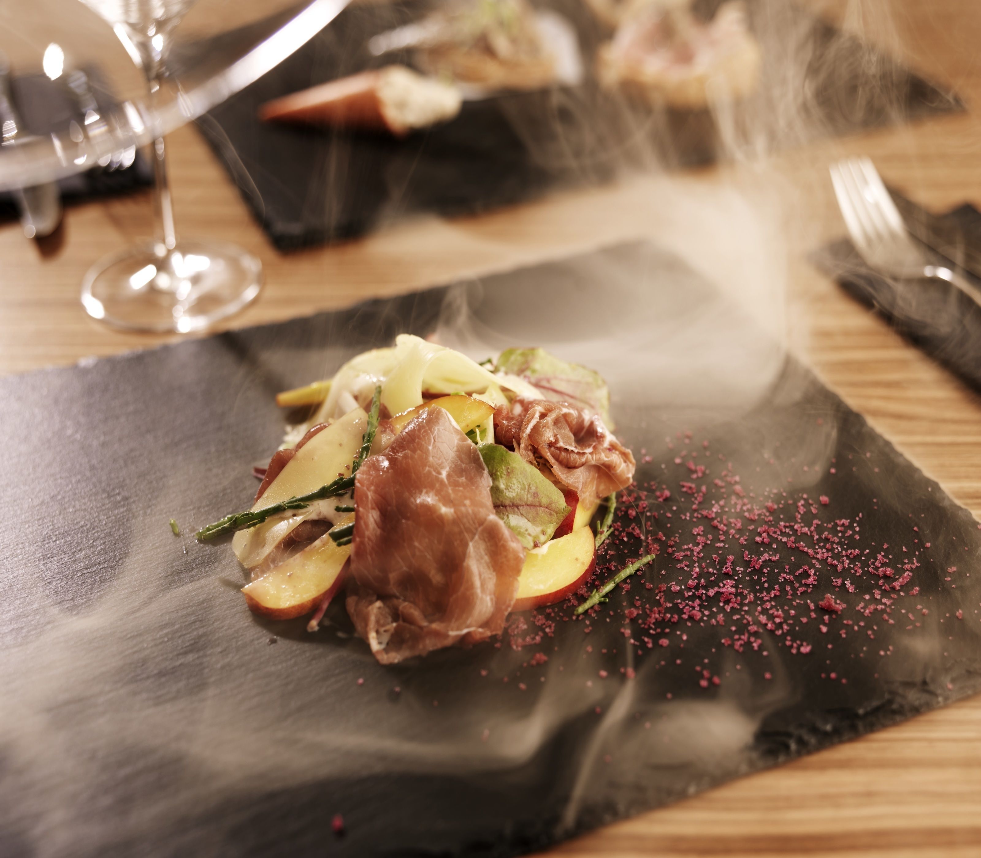 A dining table with a black slate plate containing a dish of food, a glass of wine, and a fork and knife on a napkin.