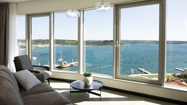 A living room with a large window overlooking a marina and water. There is a sofa, a small round table with a plant, and a chair.