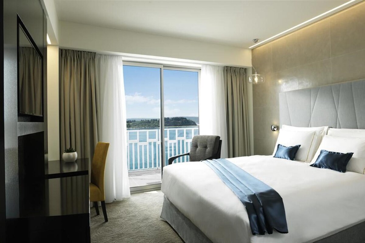 A hotel room with a large bed, a chair, a desk, and a view of the sea.