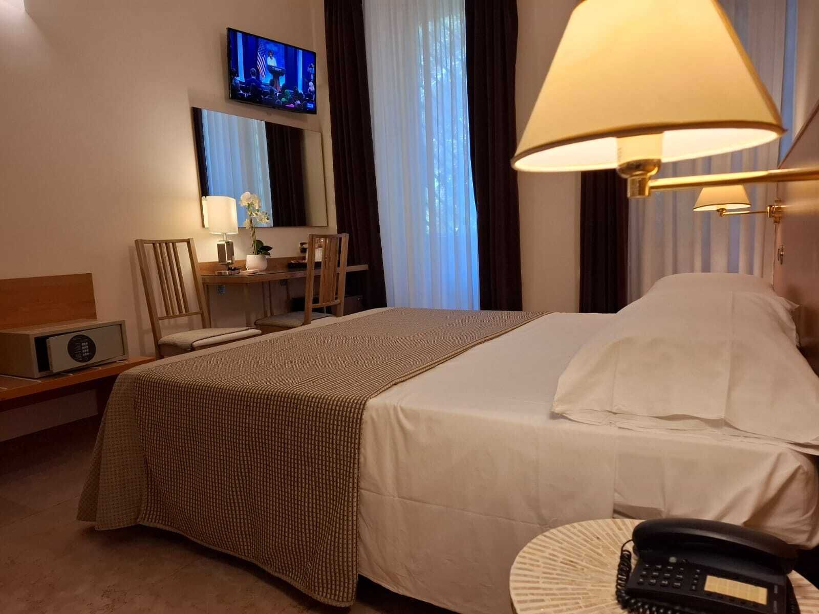Et hotelværelse med en seng, et skrivebord med lampe, et fjernsyn på væggen og en telefon på natbordet.