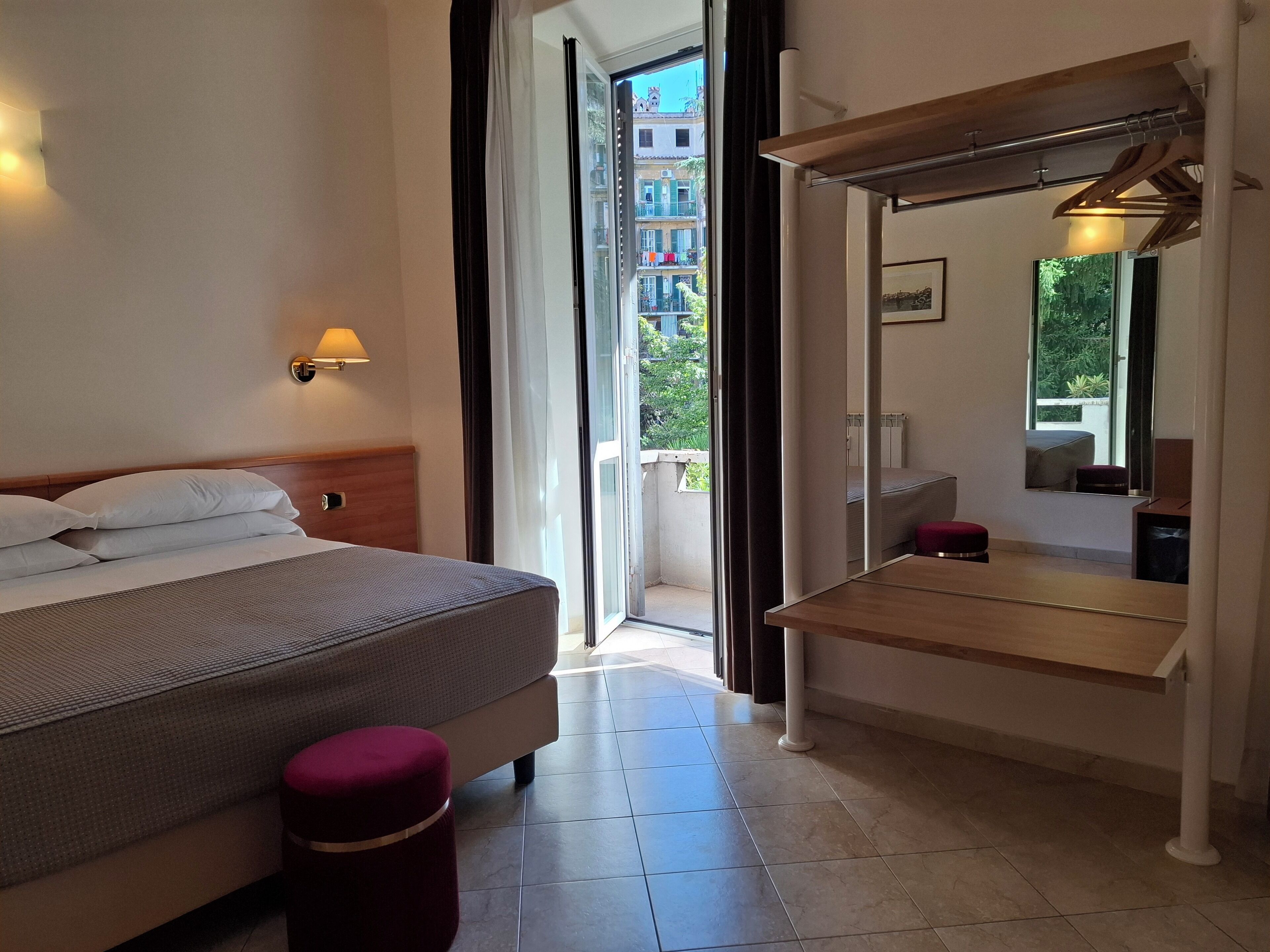 A hotel room with a bed, a bedside table, a mirror, and a view of the outside balcony.