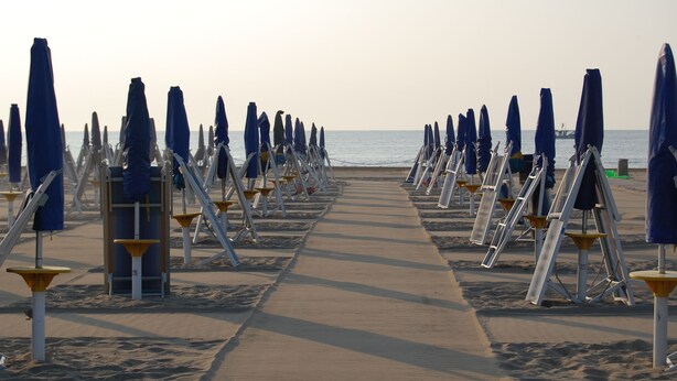 A beach with rows of blue umbrellas and white lounge chairs aligned along a concrete walkway. - Oasis Lagoon Estate (Venice)