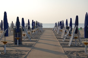 A beach with rows of sun loungers and umbrellas, a concrete walkway, and the ocean in the background.