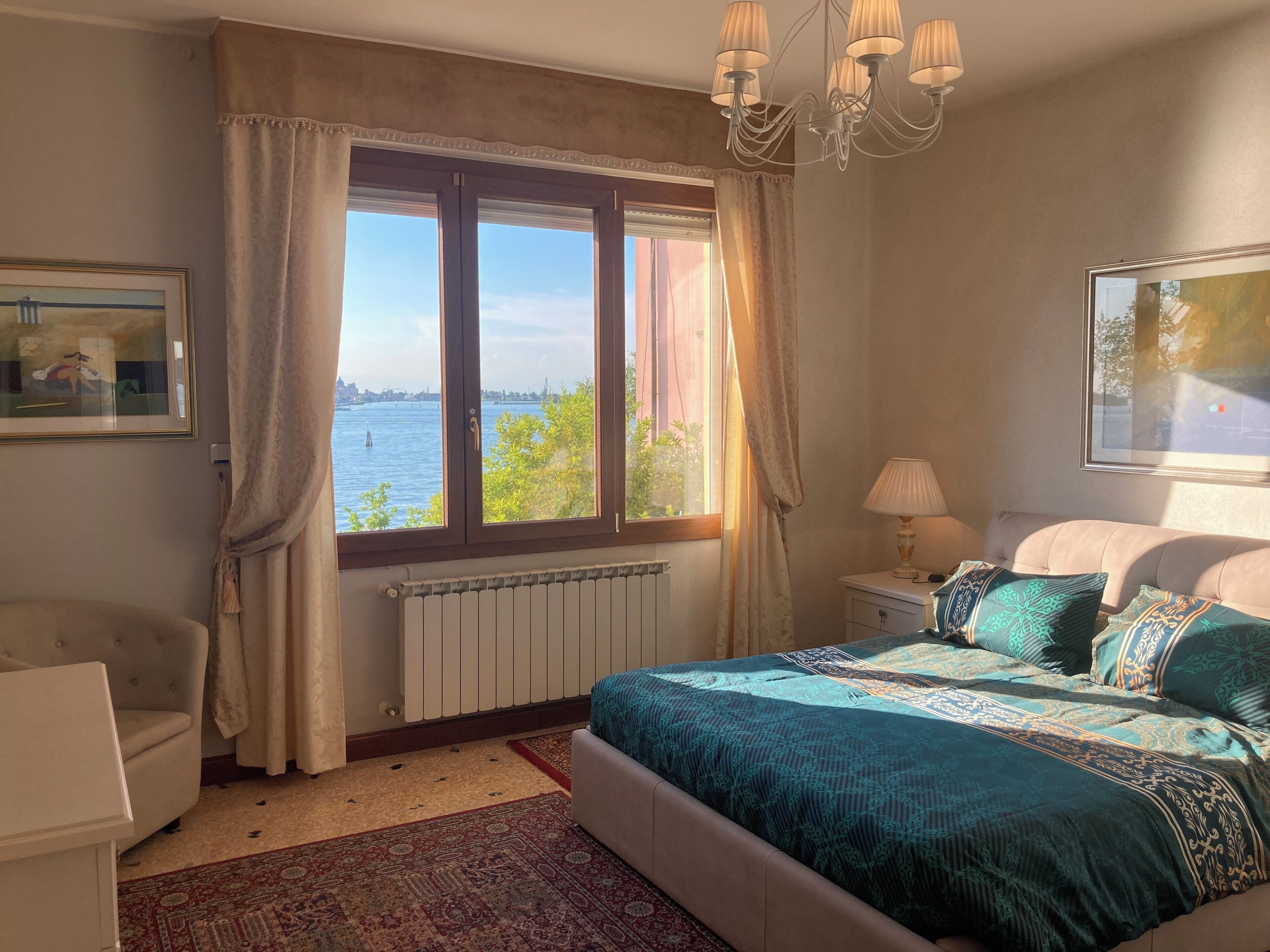Ein Schlafzimmer mit großem Fenster, einem Bett mit gemusterten Textilien, einem Heizkörper, einem Stuhl und einer Lampe.