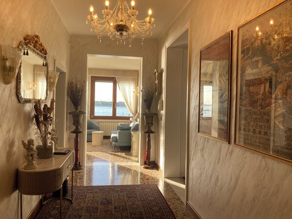 A hallway with a chandelier, a mirror, a vase with dried flowers, a framed picture, and a view of the sea through a window.