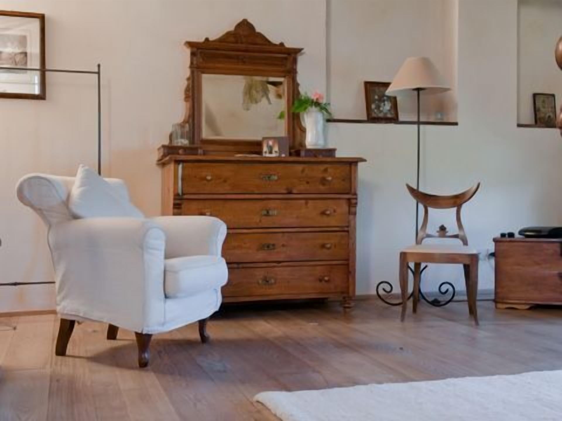 Een kamer met een witte fauteuil, een houten dressoir en een kleine tafel met een lamp.