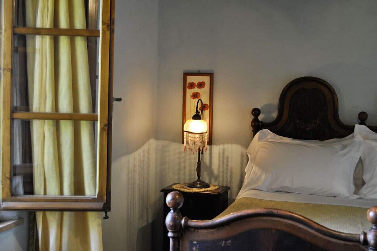 A bedroom with a wooden bed, a bedside table with a lamp, a framed picture on the wall, and a window with curtains.