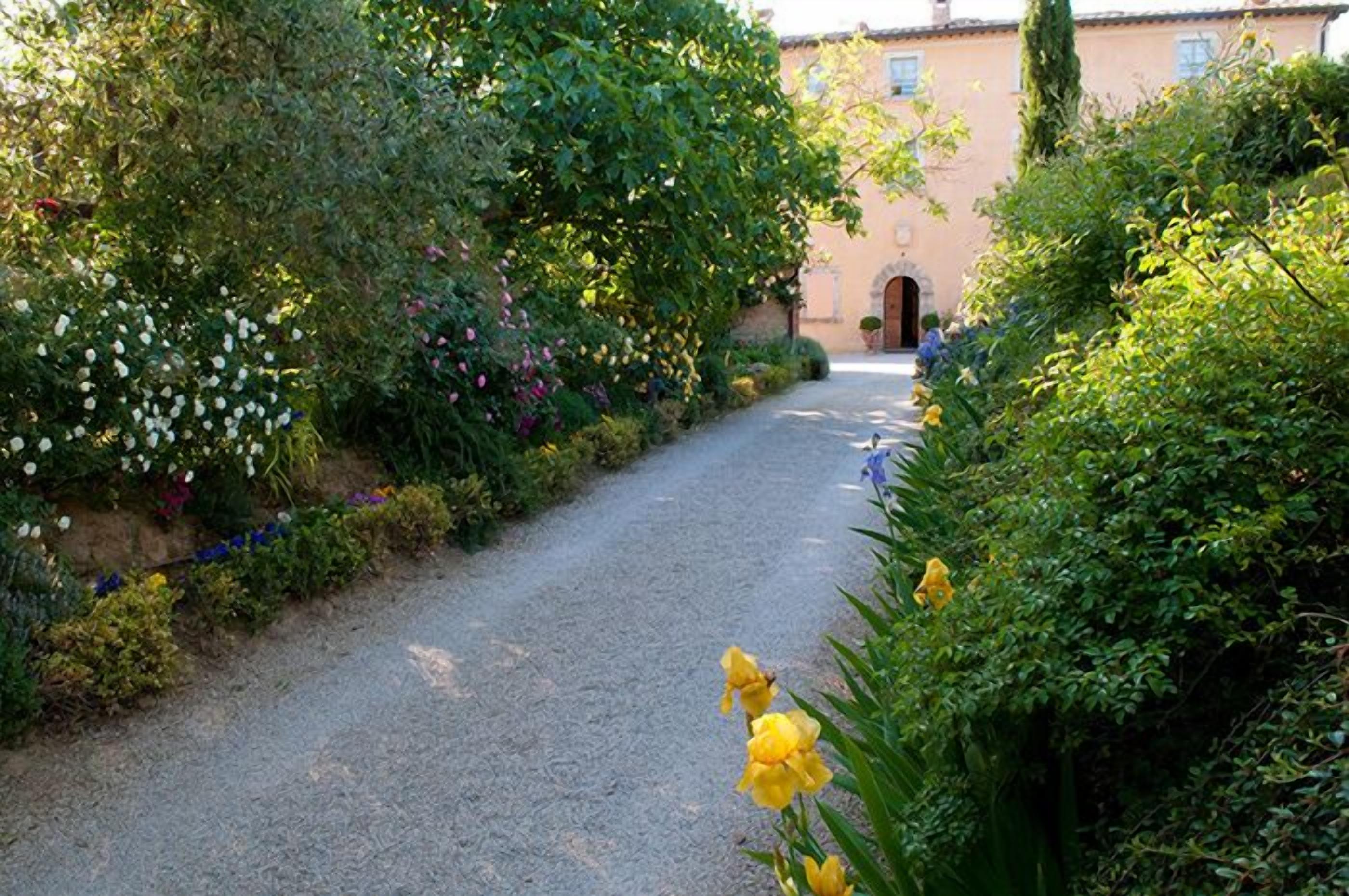 Un chemin de gravier mène à un bâtiment à la façade traditionnelle, entouré d’une végétation luxuriante et de plantes en fleurs.