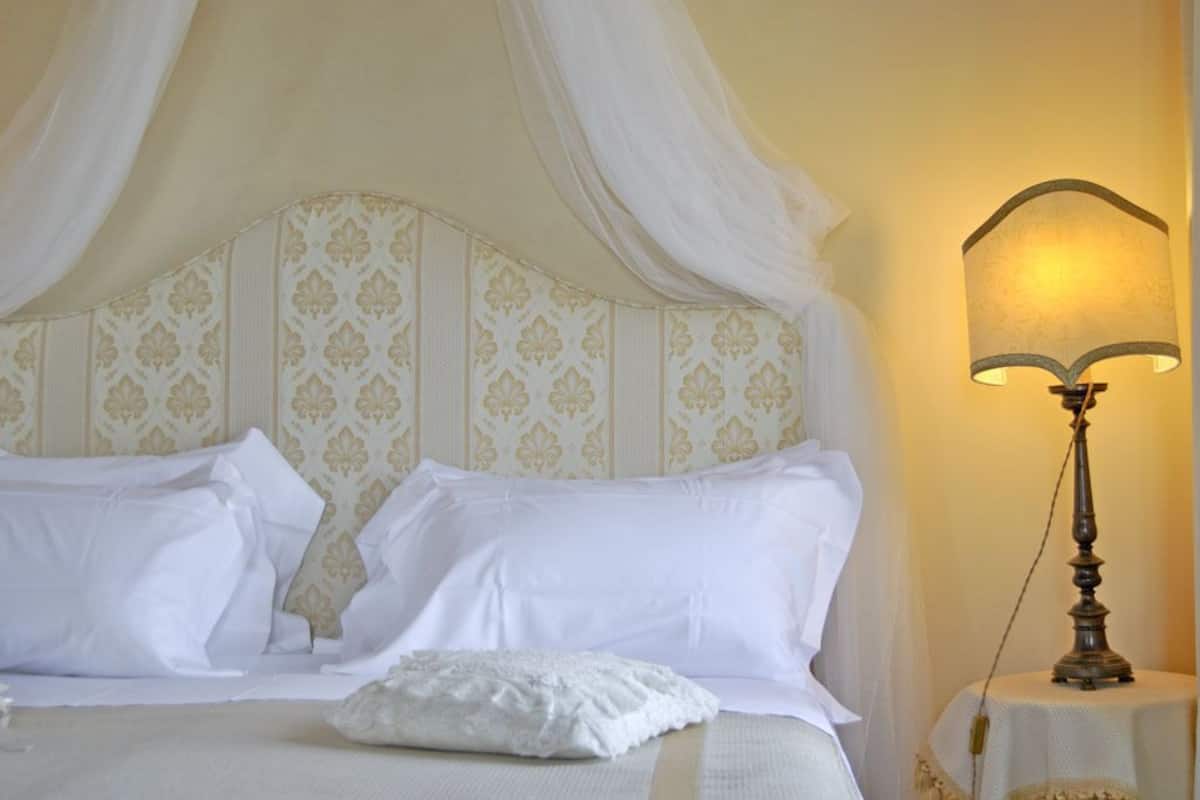 A neatly made bed with white linens and a canopy, a bedside lamp, and a patterned wallpaper.