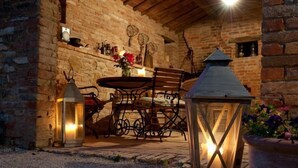 Un'area pranzo esterna rustica con pareti in pietra, un tavolo in legno, sedie in metallo e due lanterne illuminate.