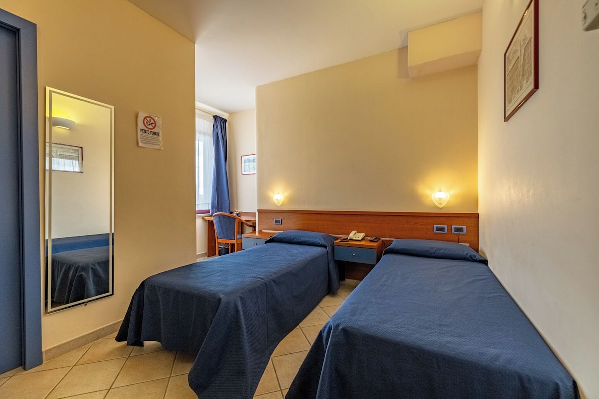 A hotel room with two single beds, a desk with a chair, a mirror, and a wall-mounted light.