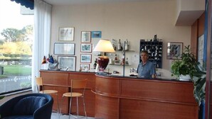 A reception area with a wooden desk, chairs, and a receptionist. There are framed pictures on the wall, a lamp, and a plant.