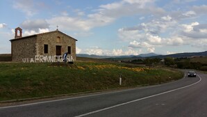 A stone building with a bell tower and a cross, labeled "VIA FRANCIGNA," on a grassy hill with a road in the foreground.