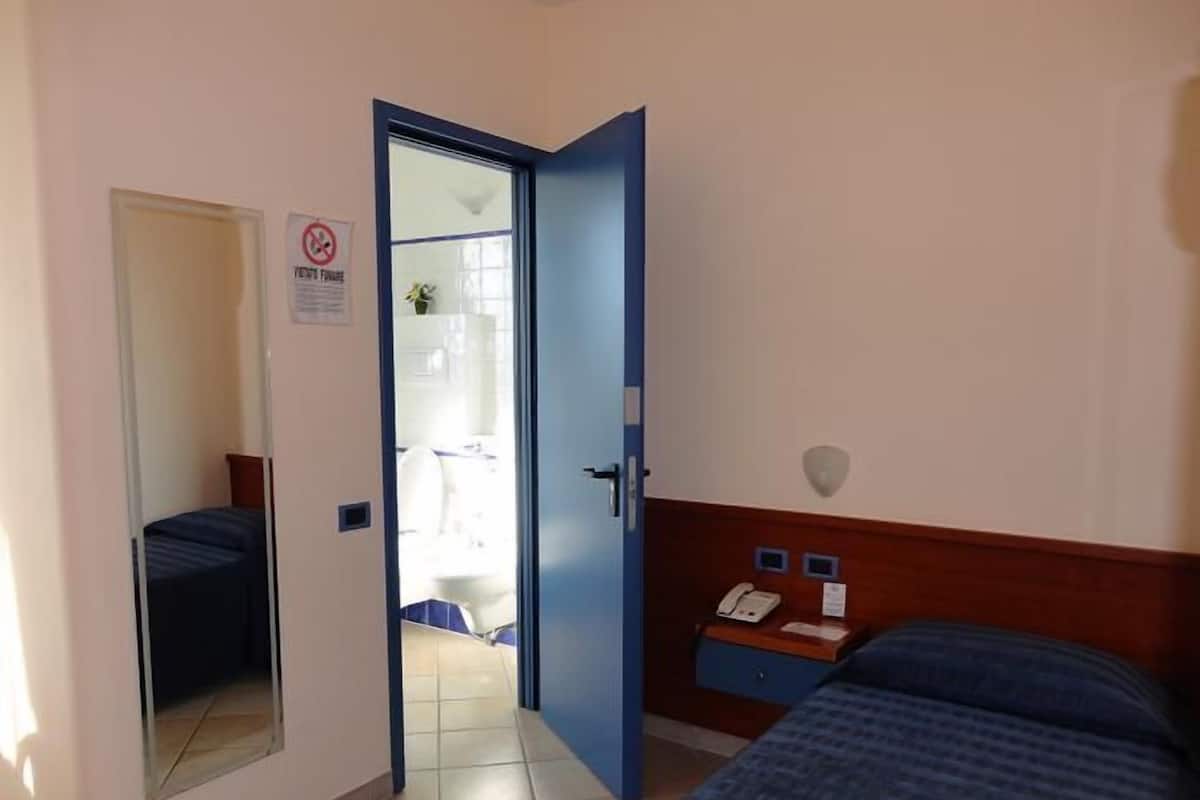 A hotel room with a bed, a bedside table with a phone, a mirror, and a door leading to a bathroom.