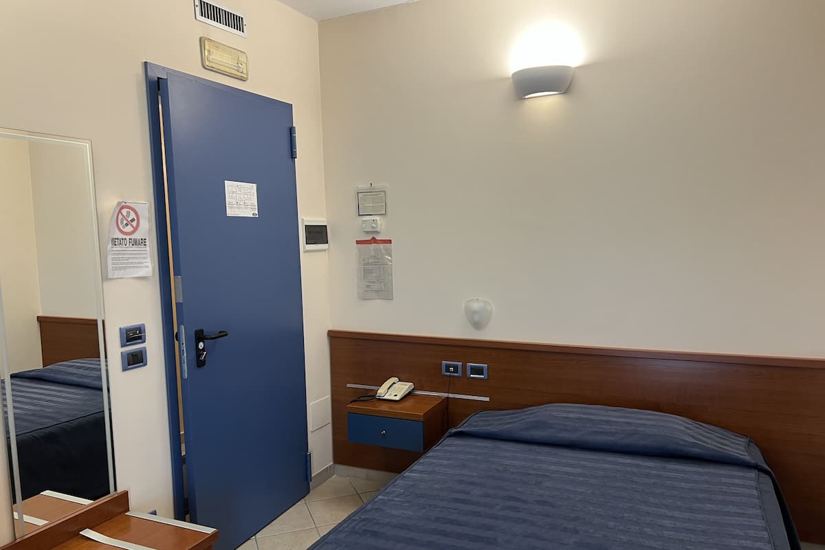 A hotel room with two beds, a wooden headboard, a bedside table, a mirror, a door with a sign, and a wall-mounted light fixture.