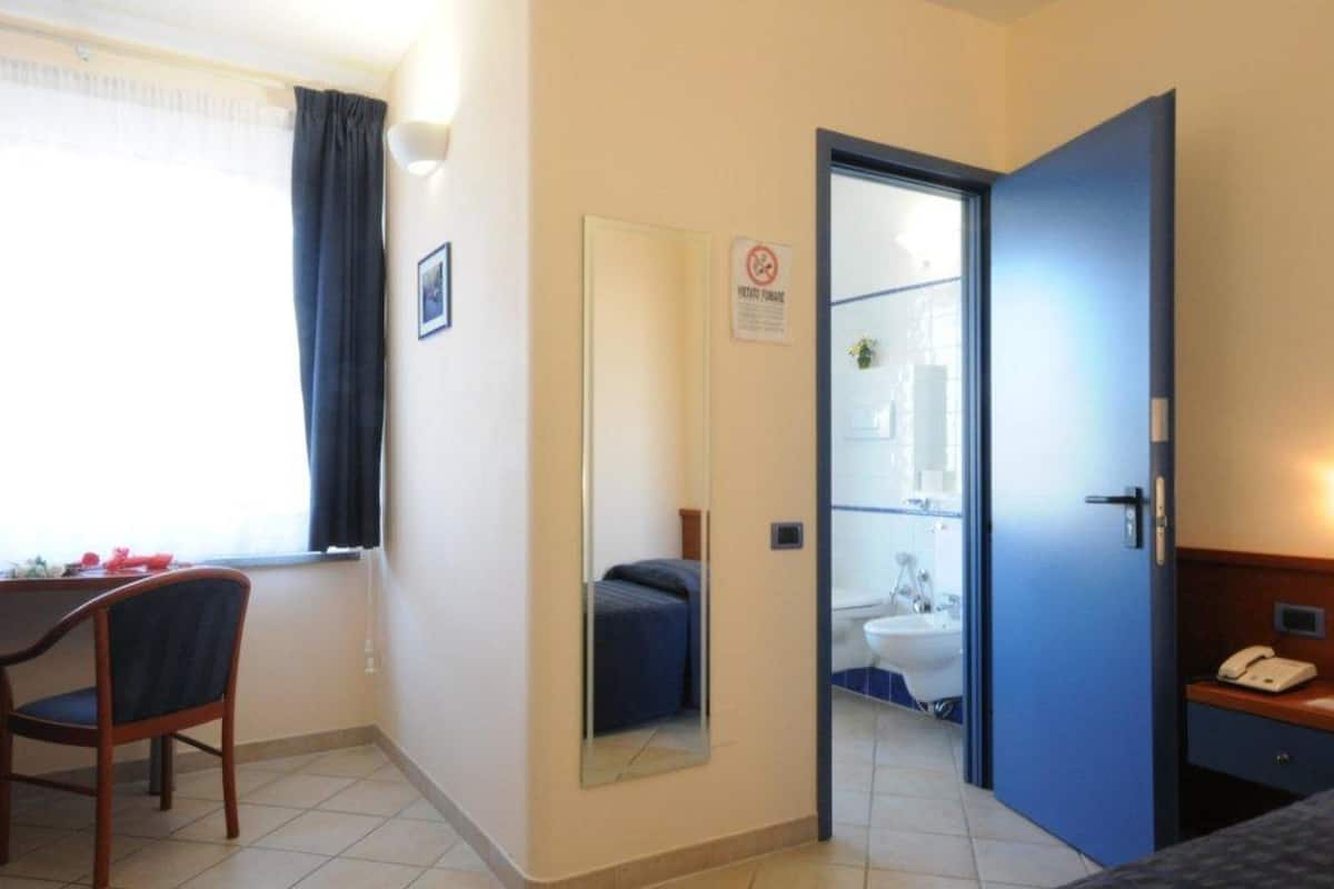 A hotel room with a desk, chair, and a mirror reflecting the bathroom.