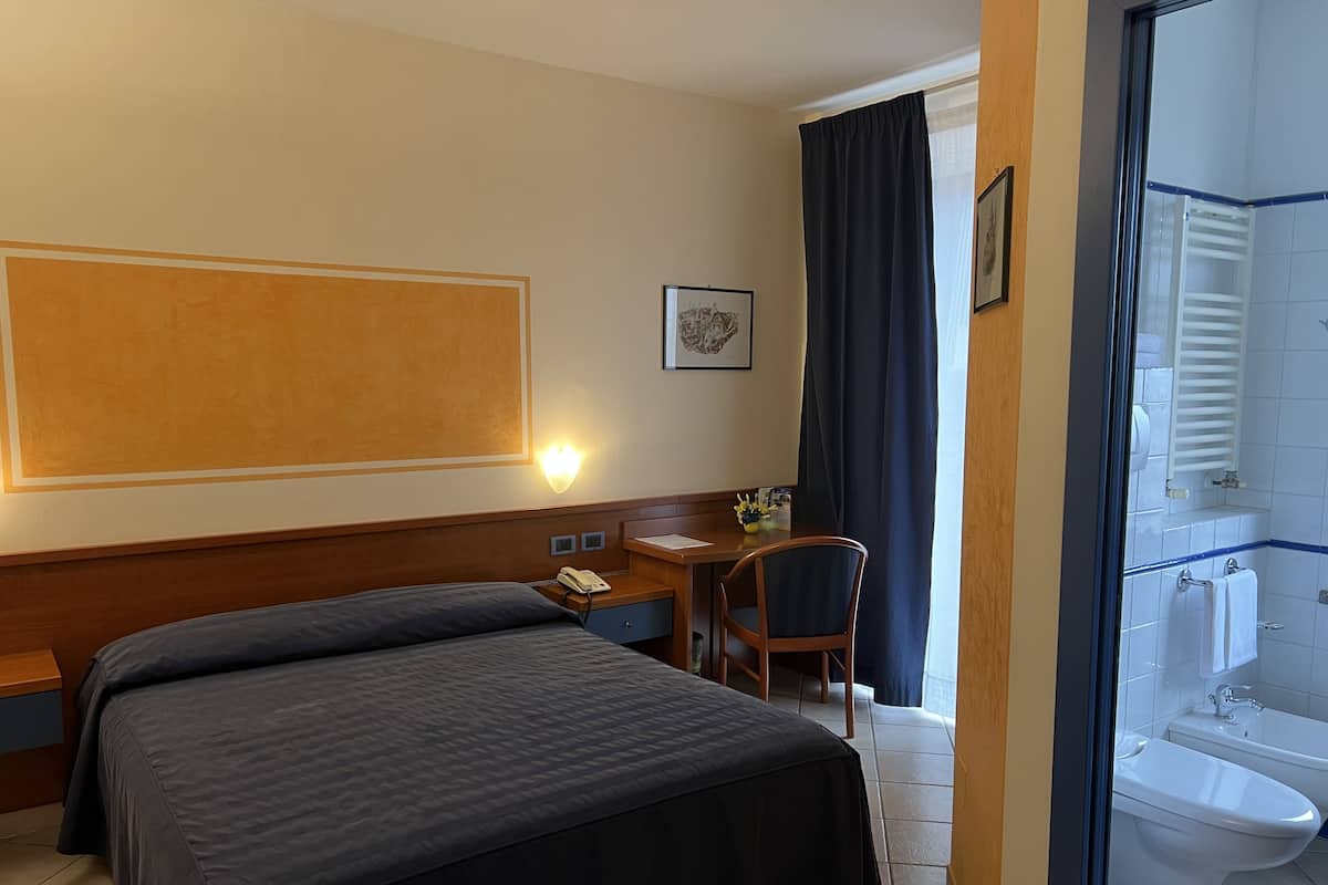 A hotel room with a large bed, a desk with a telephone, a chair, a framed picture on the wall, and a bathroom with a toilet and sink.