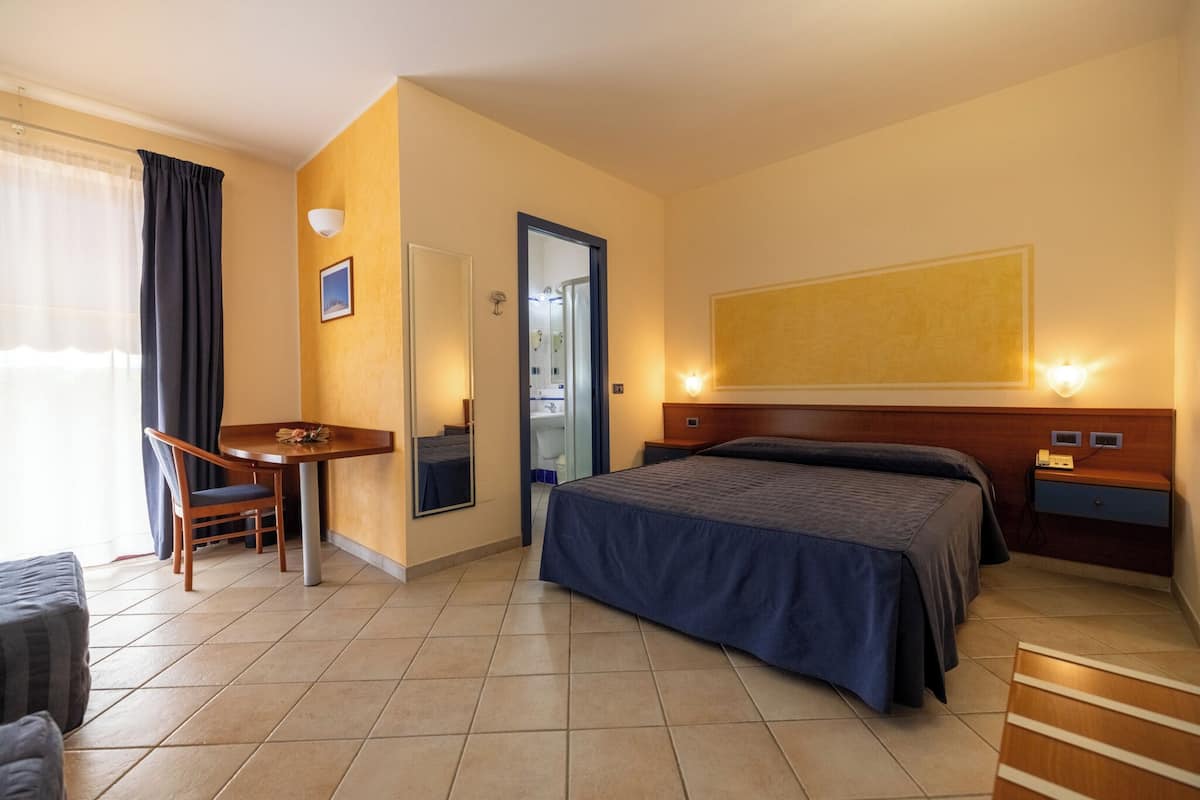 A hotel room with a bed, a desk, a chair, a small table, and a large painting on the wall.