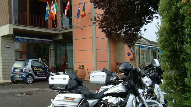A motorcycle parked in front of a building with multiple flags, a car, and a sign that reads "Hotel".