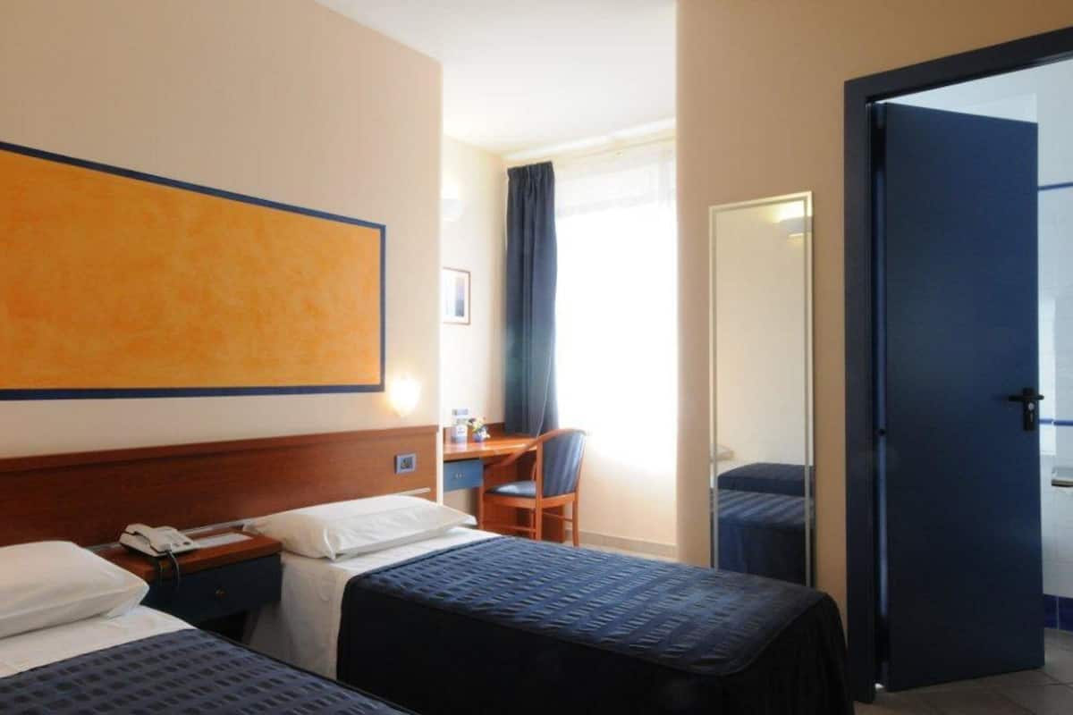 A hotel room with two beds, a desk, a chair, and a large window with curtains.