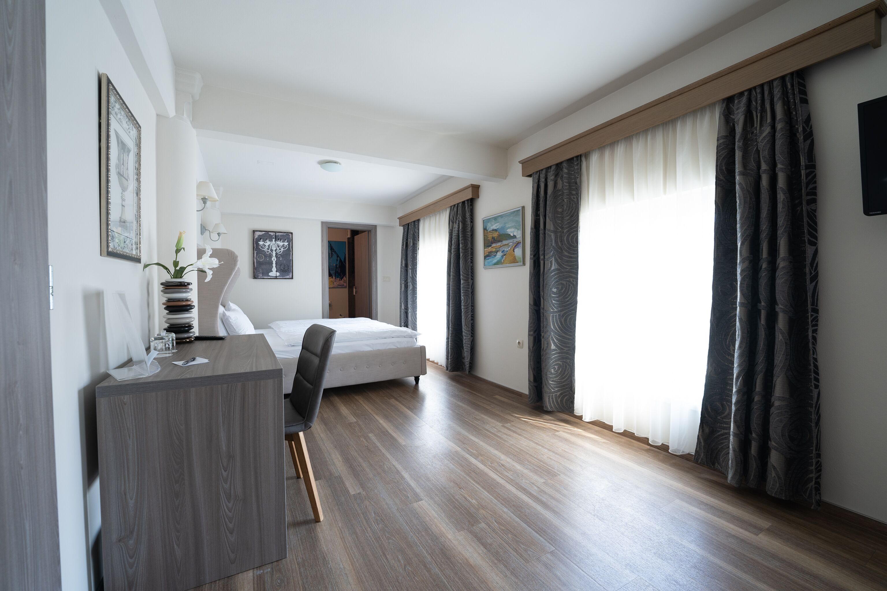 Una habitación de hotel moderna con suelo de madera, una cama, un escritorio con silla y ventanas amplias con cortinas.