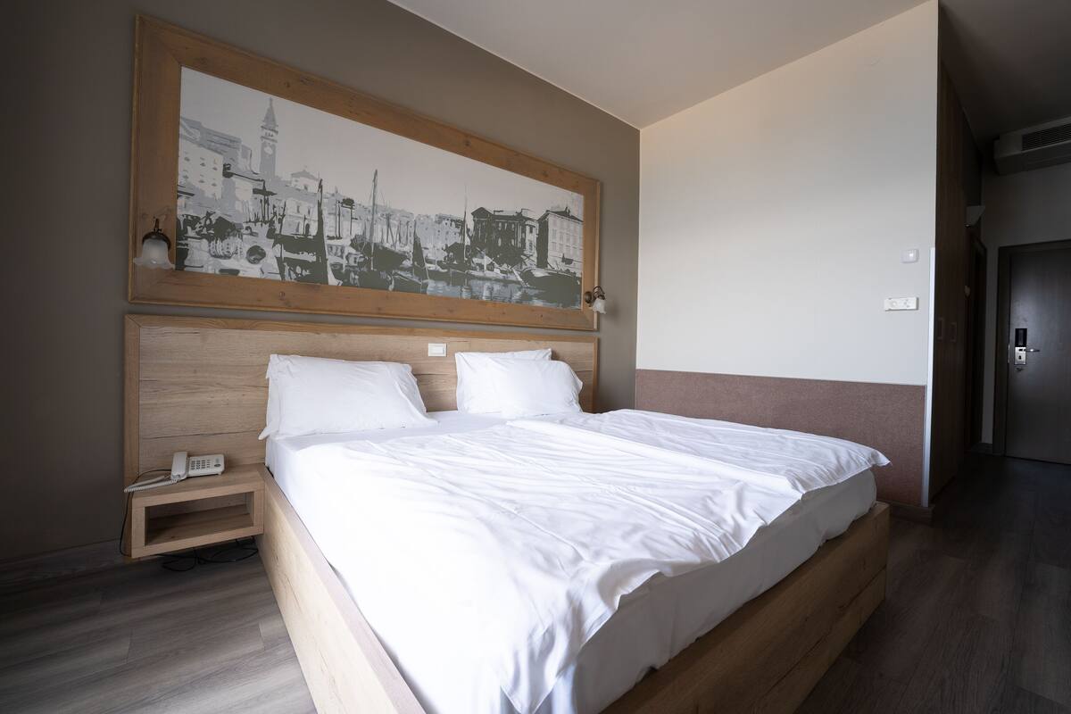 A hotel room with a large bed, a bedside table with a phone, and a wall-mounted artwork depicting a cityscape.
