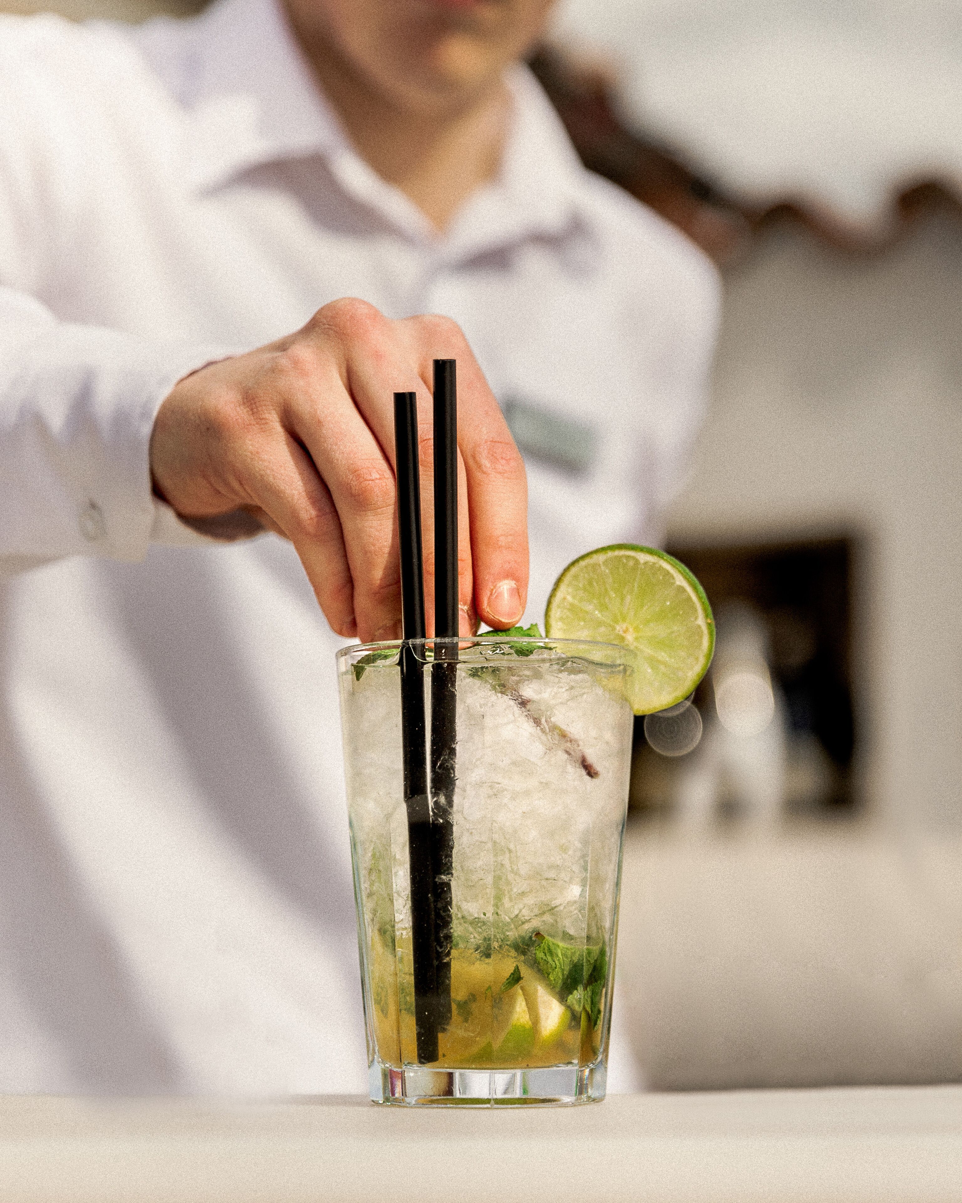 Eine Person in einem weißen Hemd hält ein Getränk mit schwarzen Strohhalmen, einer Limettenscheibe und Minzblättern im Glas.