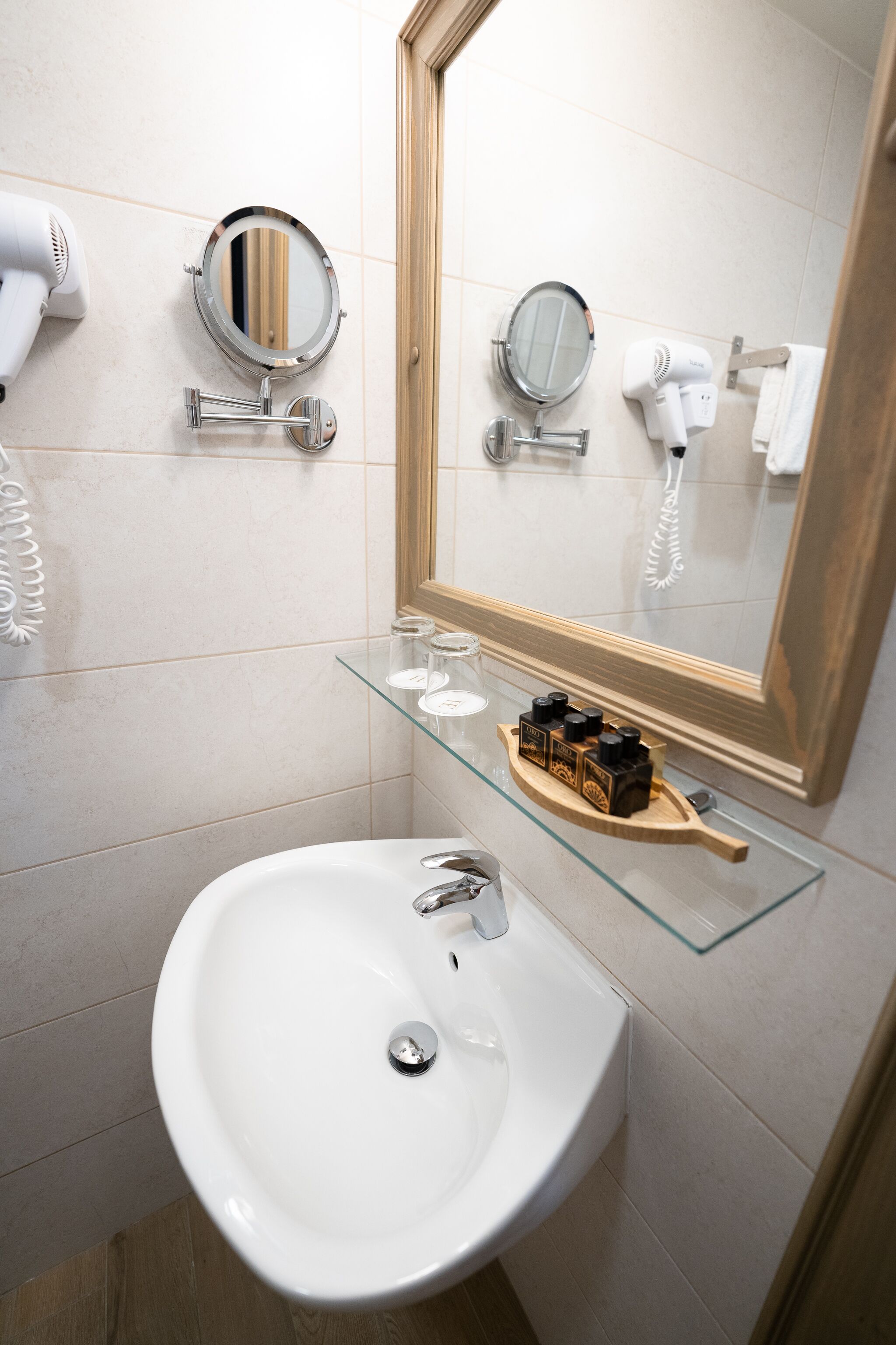 A bathroom with a white sink, two round mirrors, a glass shelf with toiletries, and a towel rack.