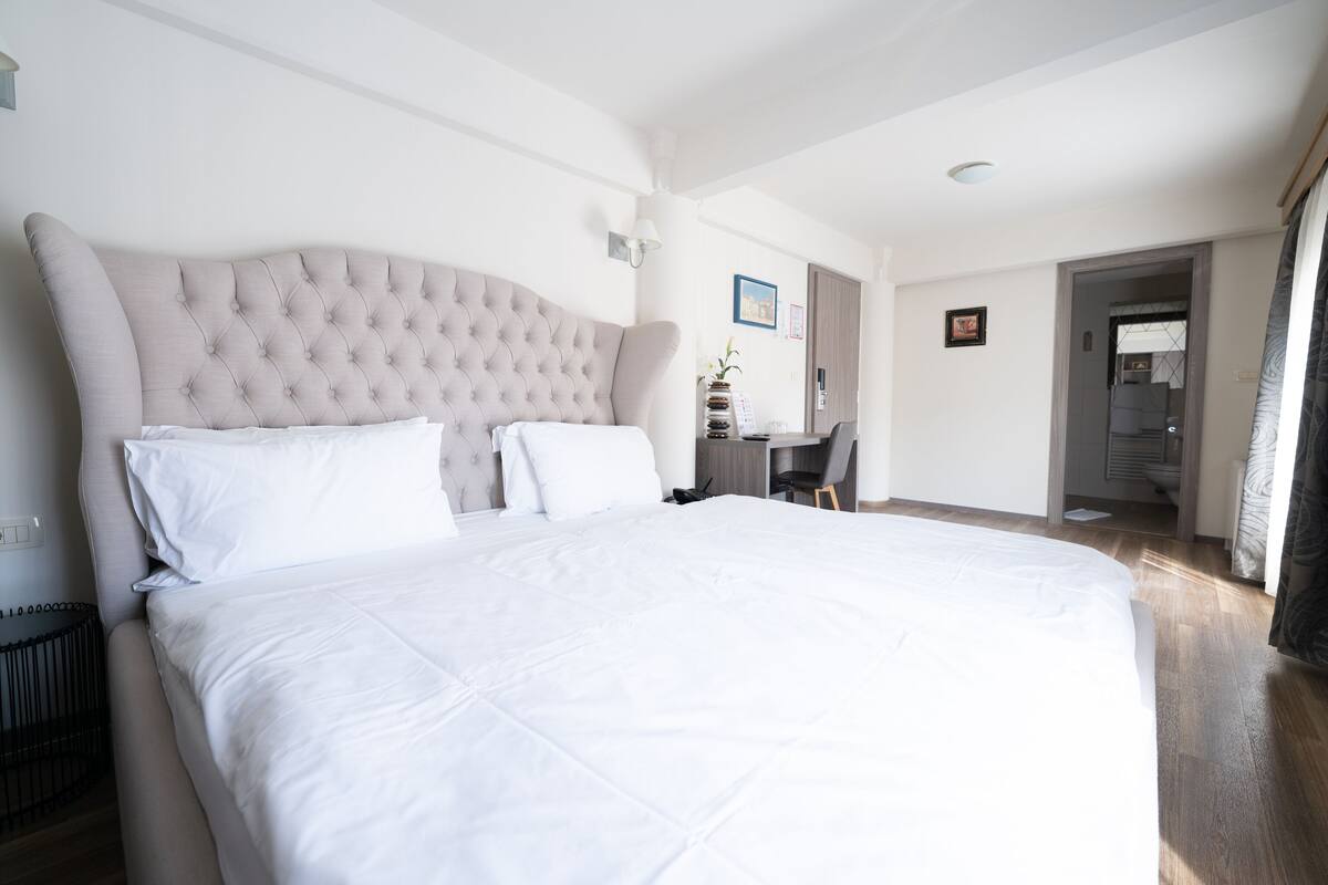 A large bed with a tufted headboard, a bedside table, and a chair in a well-lit room with wooden flooring.