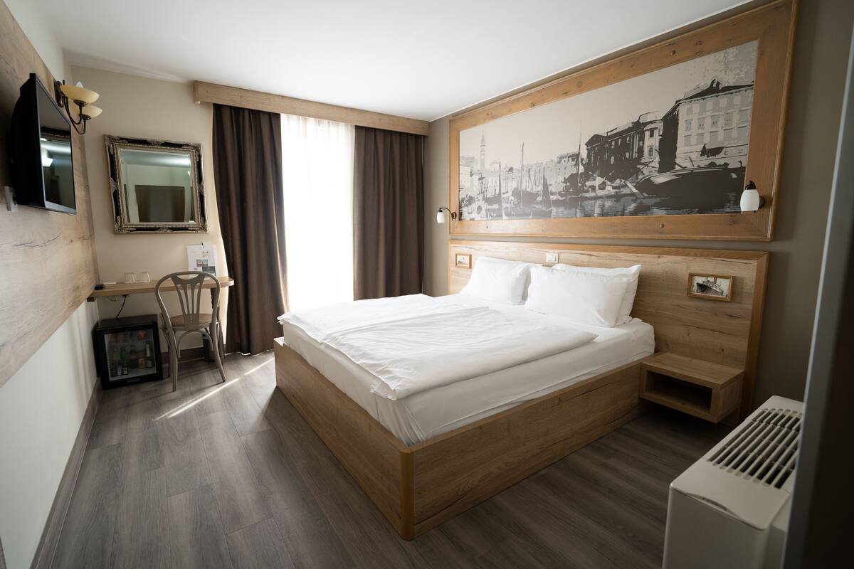 A hotel room with a wooden bed, a desk with a chair, a large painting on the wall, and a window with curtains.