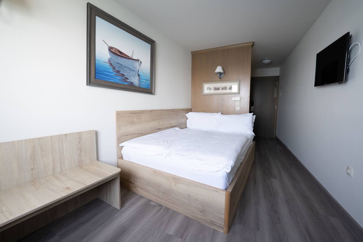 A hotel room with a wooden bed, a bench, a painting of a boat, and a TV on the wall.