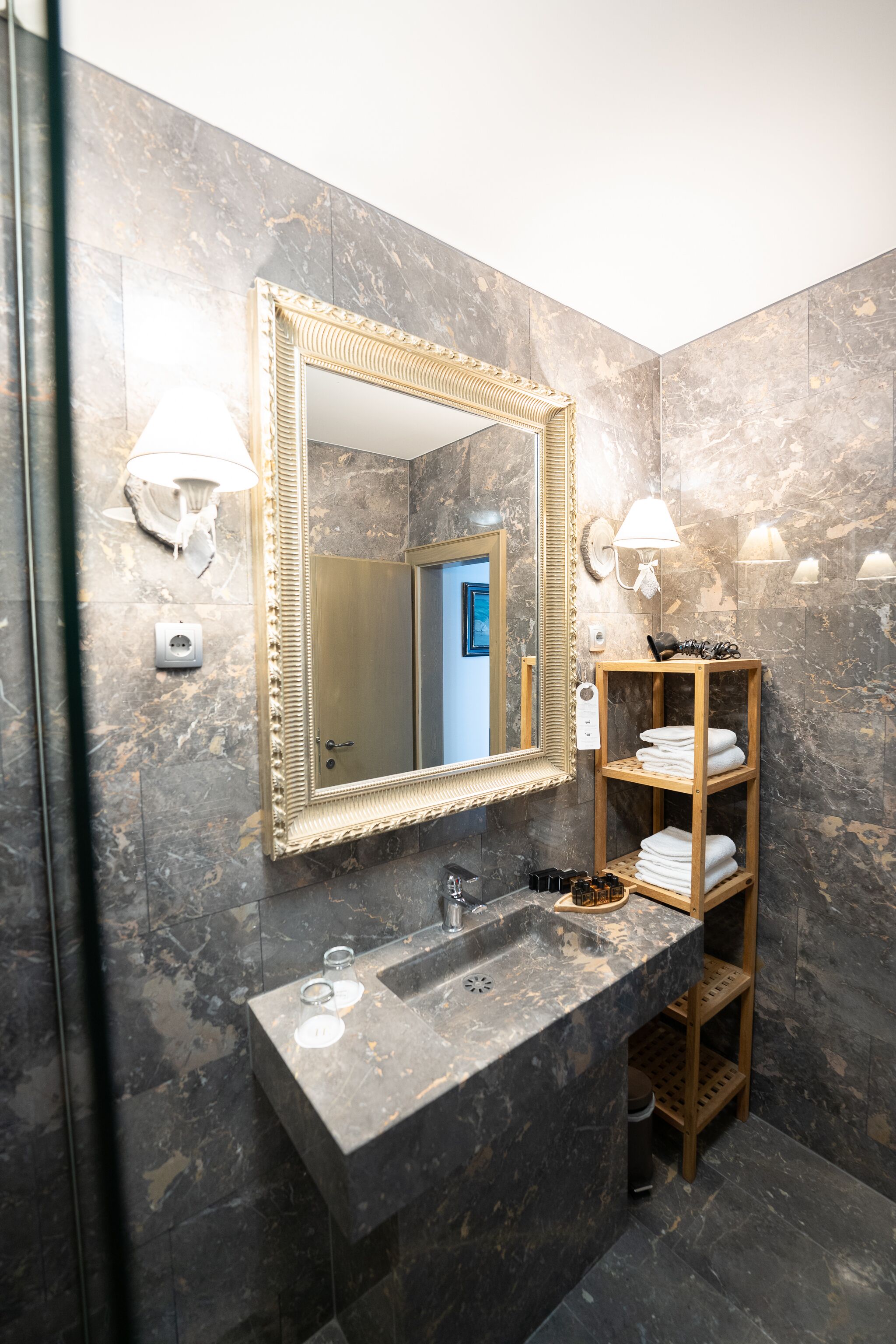 Un baño con pared de mármol, un espejo grande, repisas de madera con toallas y un lavamanos con grifo.