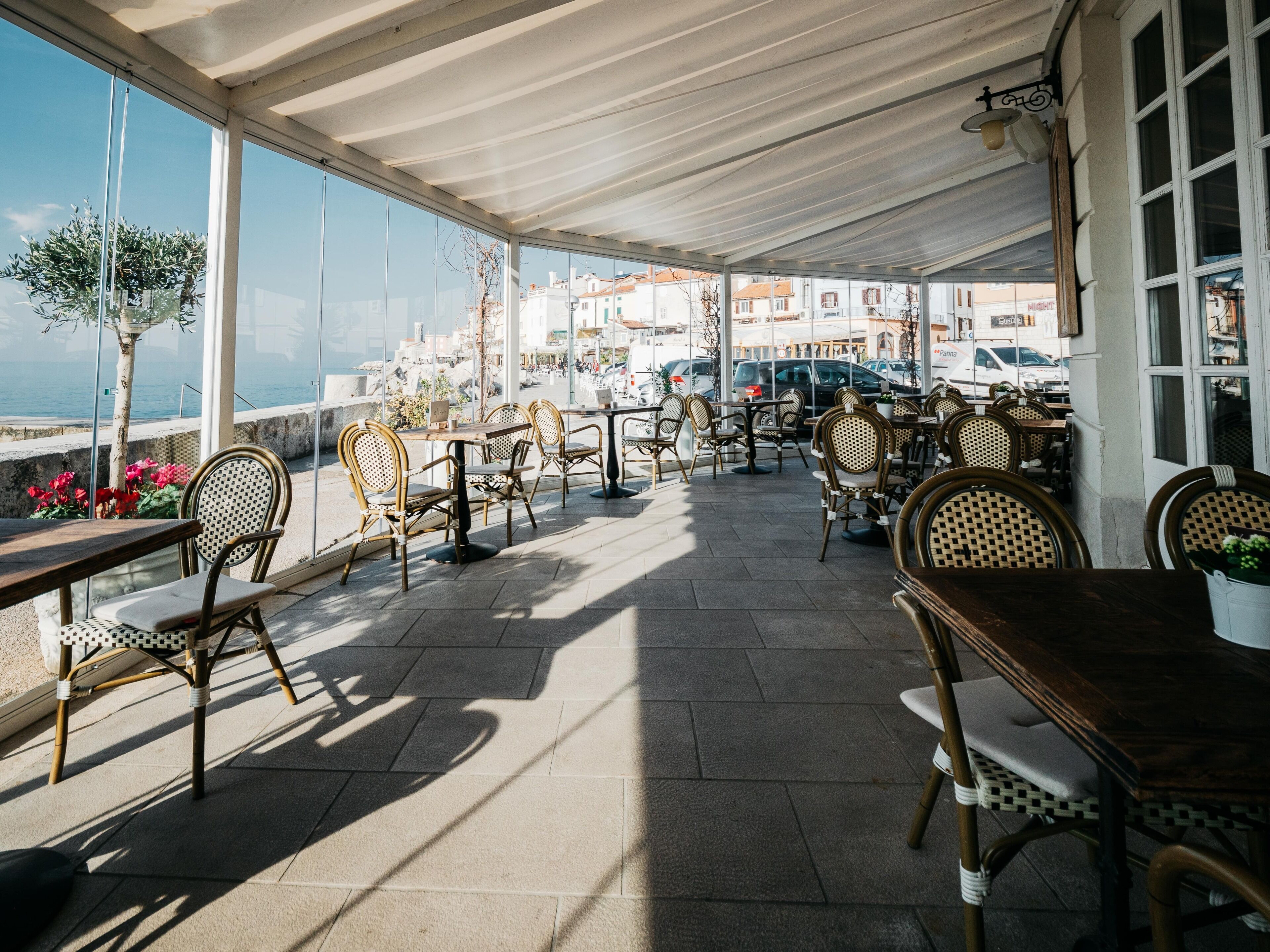An outdoor cafe with tables and chairs, overlooking a coastal area with buildings and parked cars.
