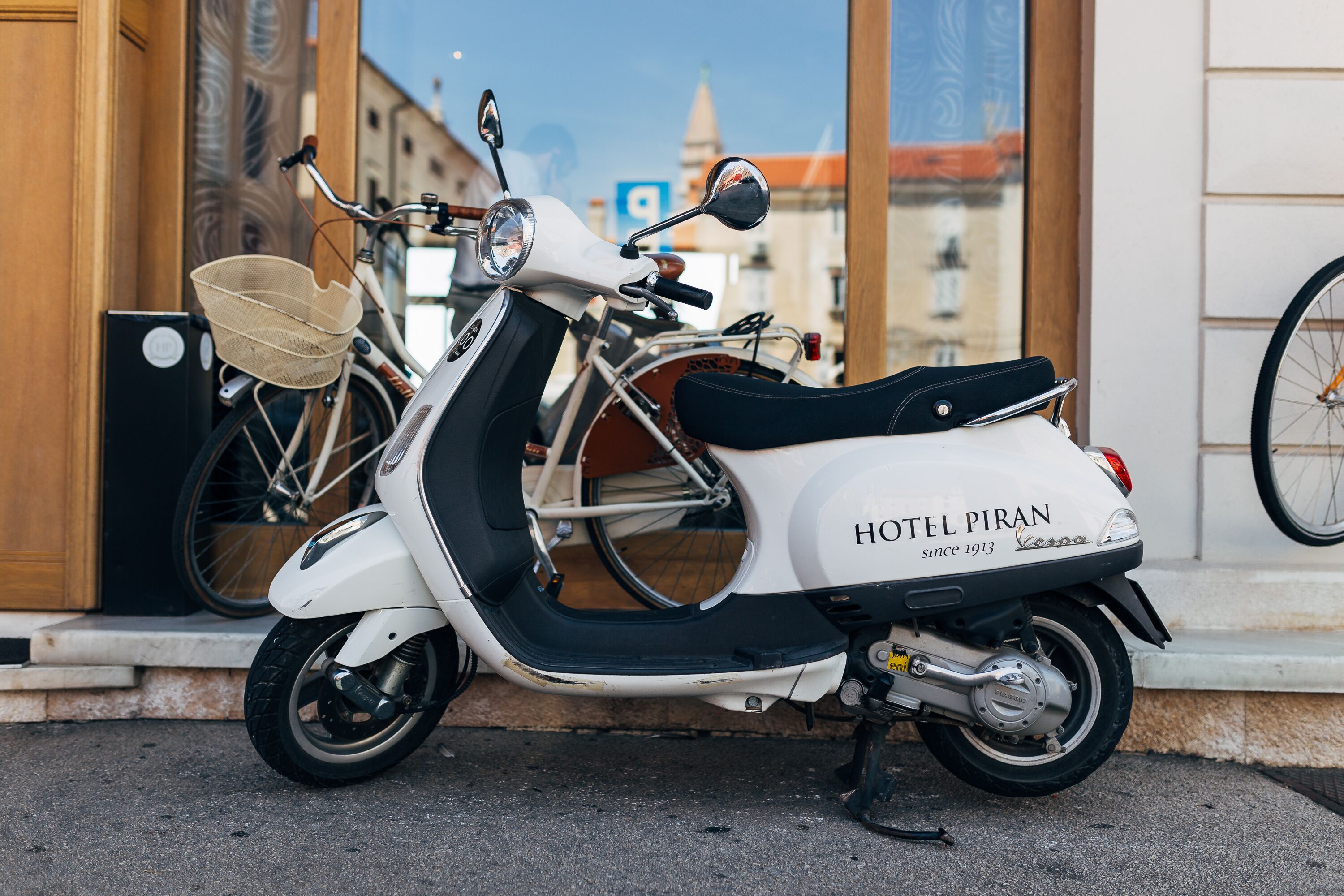 Uno scooter bianco con un cesto e un sedile nero, parcheggiato di fronte a un edificio con un cartello che recita "HOTEL PIRAN since 1913".