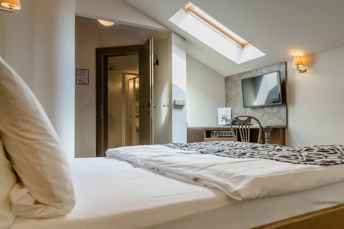 A bedroom with a bed, a desk with a computer, a chair, a TV mounted on the wall, and a skylight.