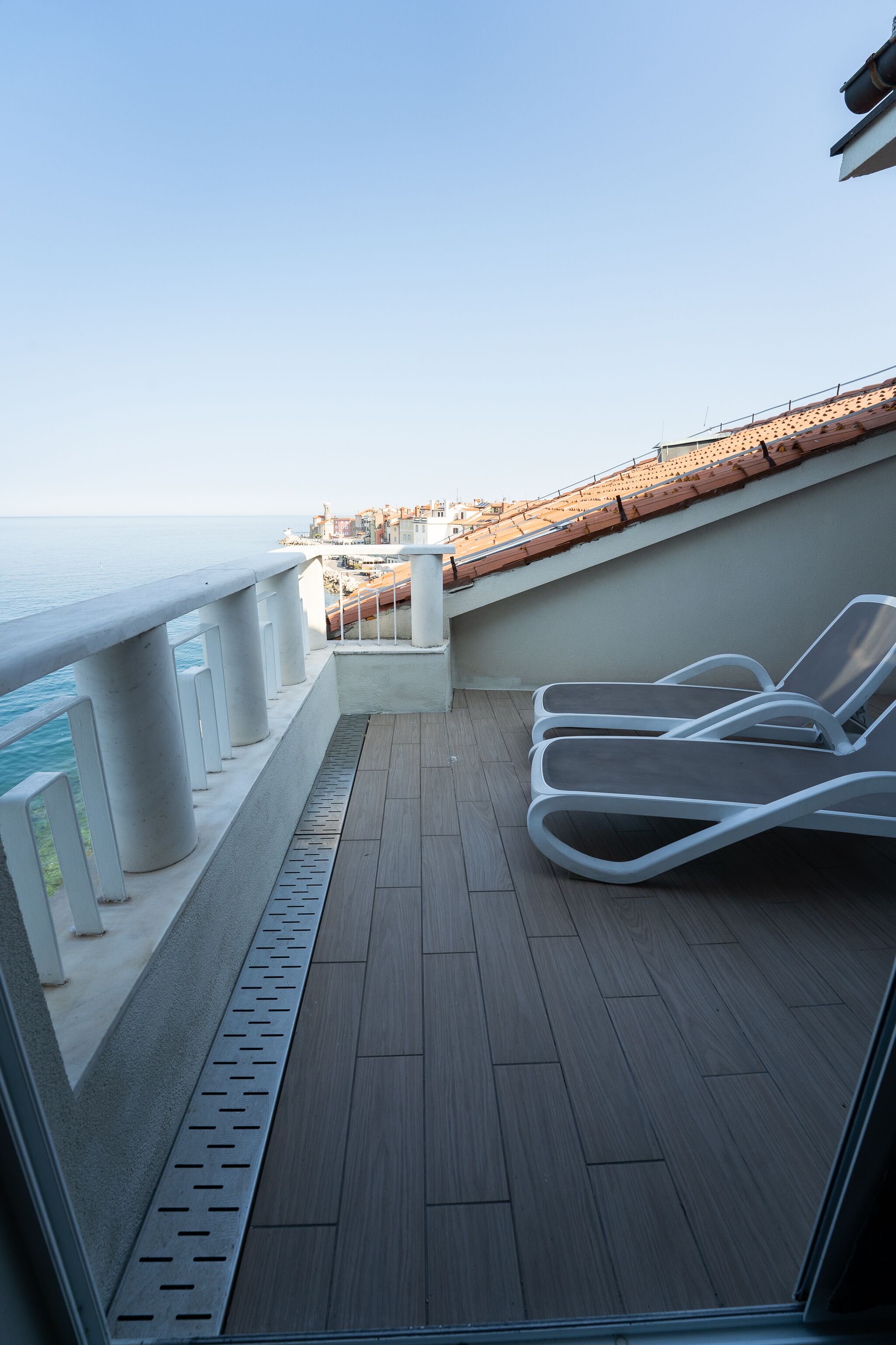 A balcony with a view of the sea, white lounge chairs, and a tiled floor.