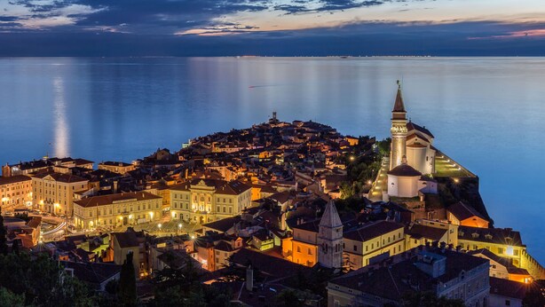 A coastal town at dusk with a prominent church tower and calm sea. - Hotel Piran (Piran)