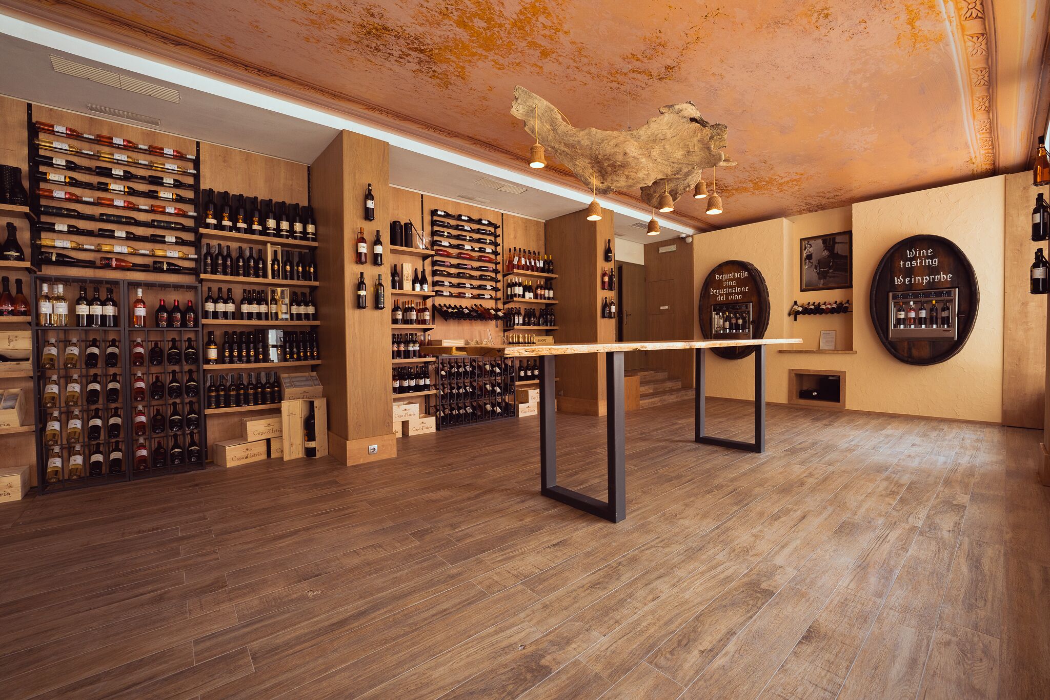 Une cave à vin avec des étagères en bois et une table centrale, où sont exposées différentes bouteilles de vin, le tout éclairé par un luminaire suspendu.