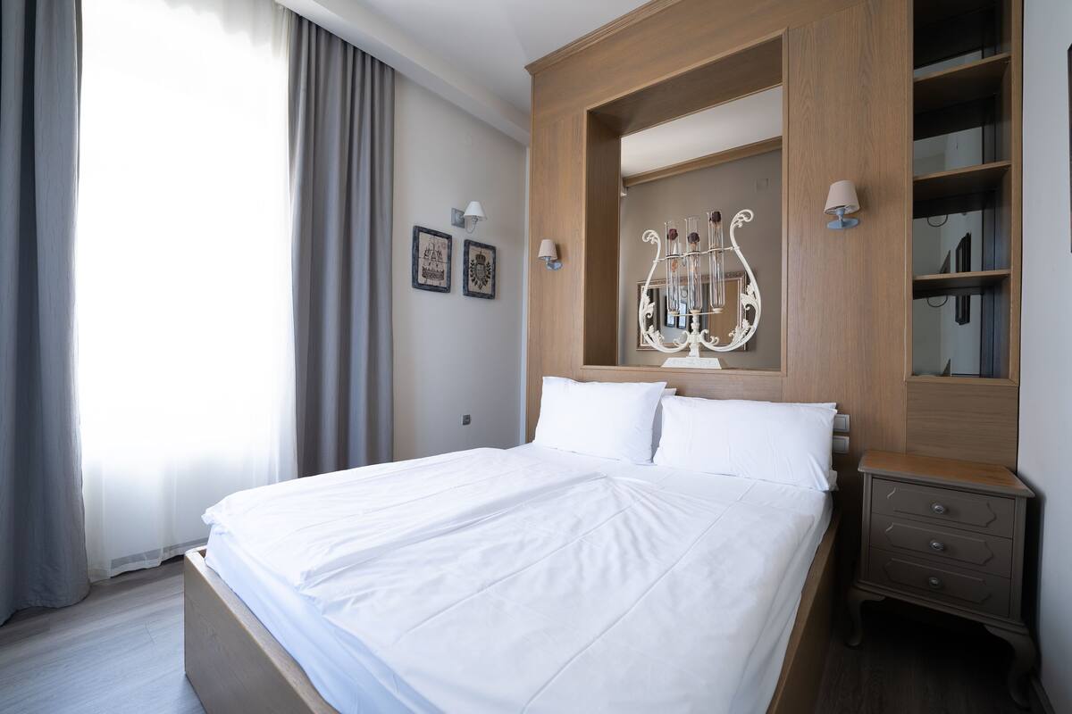 A neatly made bed with white linens, a wooden nightstand with drawers, and a wall-mounted mirror with a decorative frame.