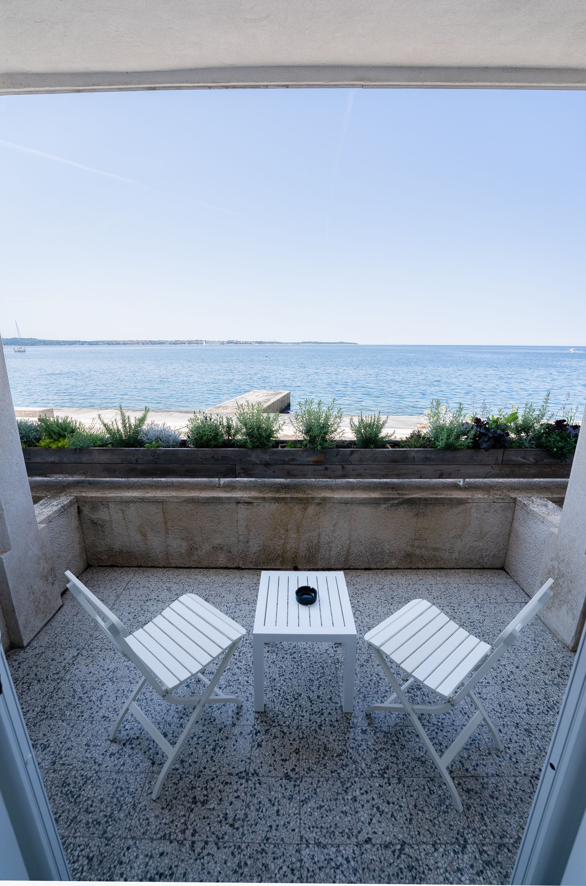 A balcony with two white chairs and a table, overlooking a body of water and a distant shoreline.
