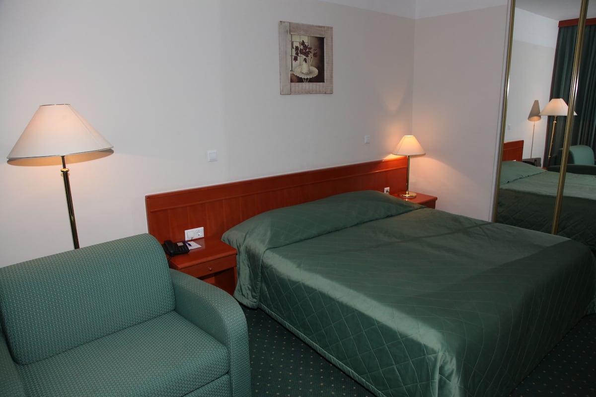 A hotel room with a bed, a chair, a nightstand, a lamp, a mirror, and a painting on the wall.