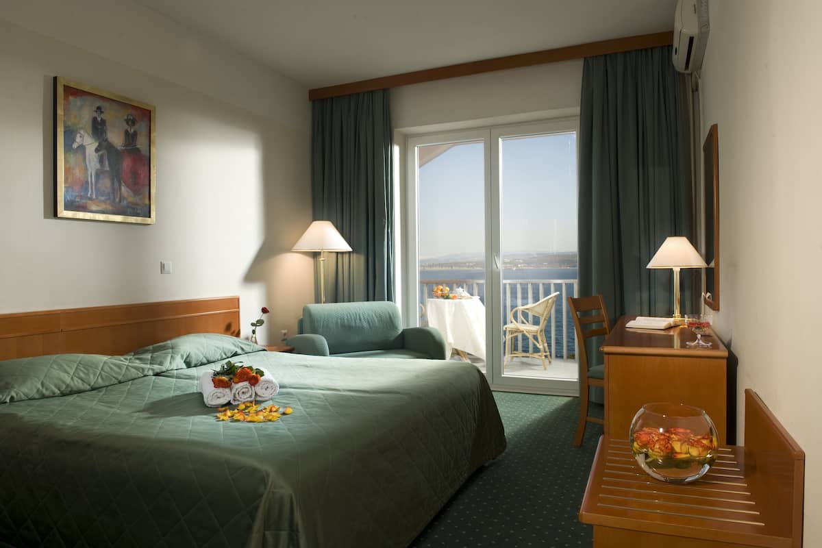 A hotel room with a bed, two chairs, a table with a vase, a painting on the wall, and a balcony with a view.