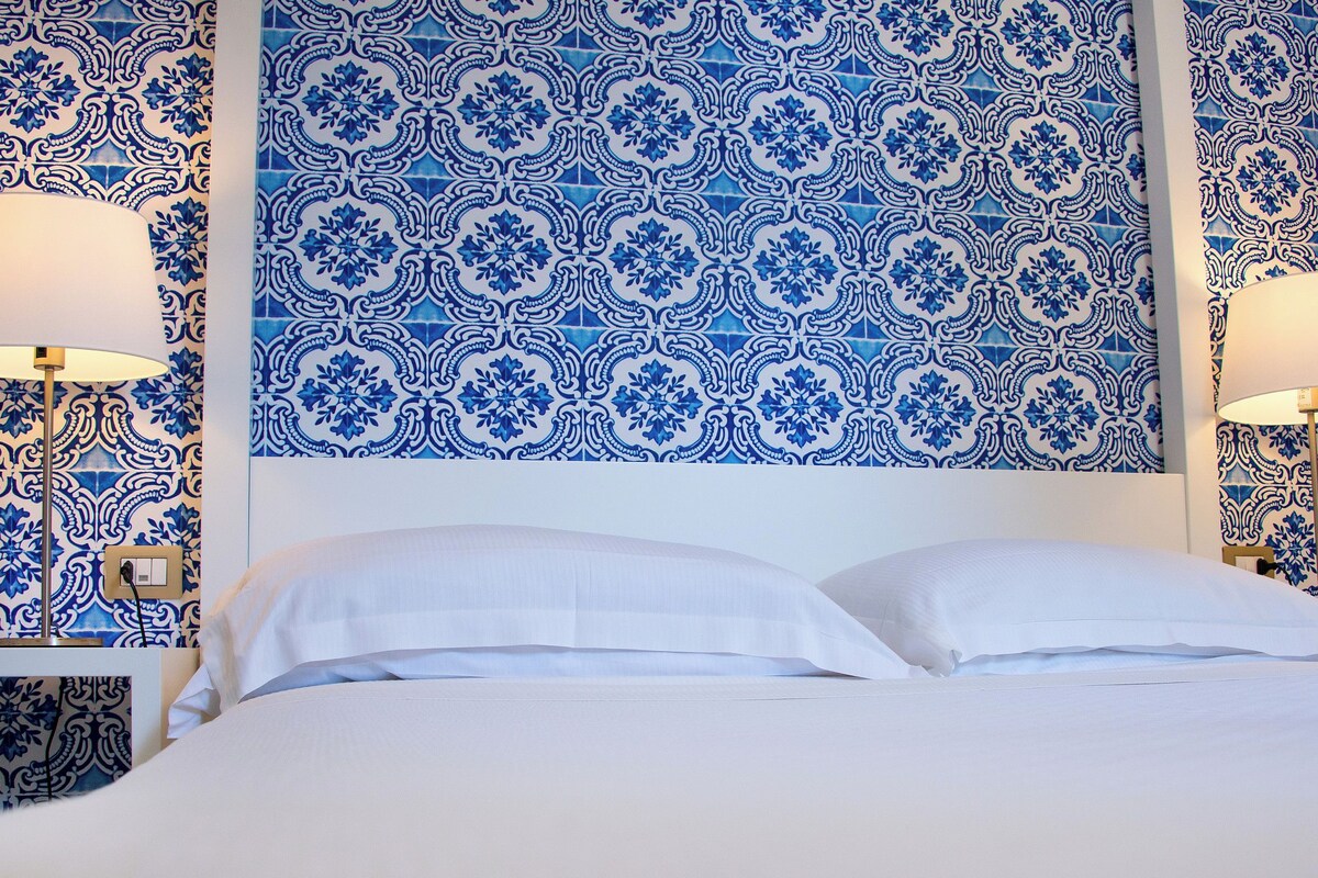 Habitación de hotel con pared de azulejos blancos y azules con diseño, dos lámparas de noche y una cama bien hecha con sábanas blancas.