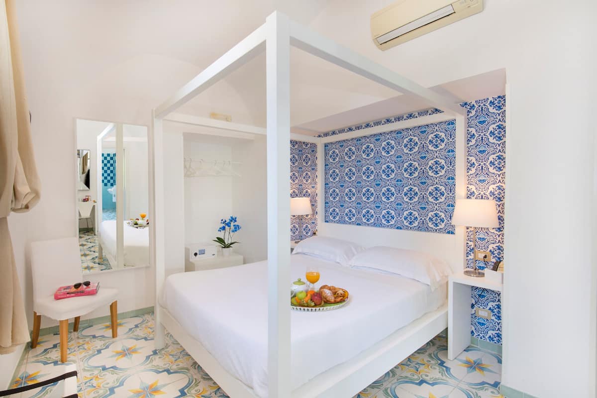 A four-poster bed with a tray of fruit and orange juice, a white nightstand, a mirror, and a bathroom with blue and white patterned tiles.