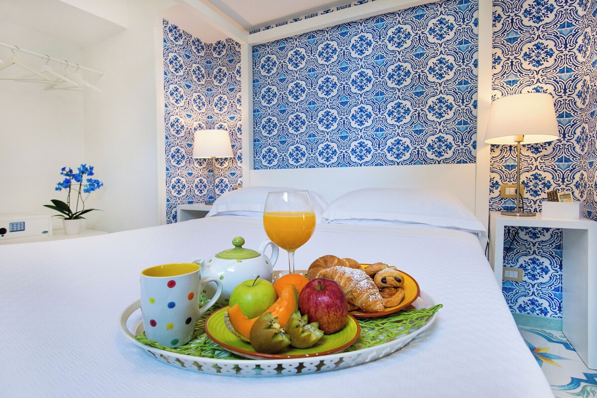 Una bandeja de desayuno ordenada con frutas, un croissant y un vaso de jugo de naranja sobre una cama en una habitación con papel tapiz azul estampado y ropa de cama blanca.