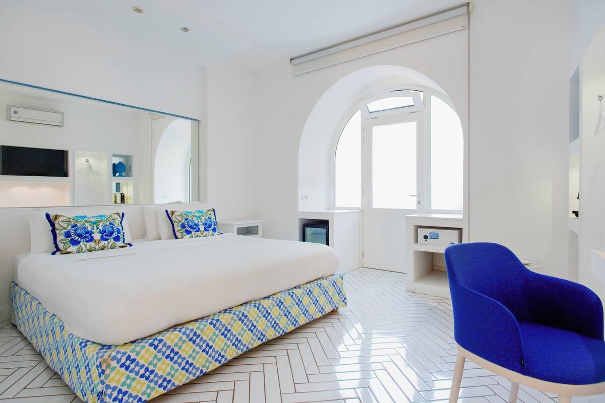 A modern hotel room with a large bed, a blue armchair, a desk, and a large arched window.