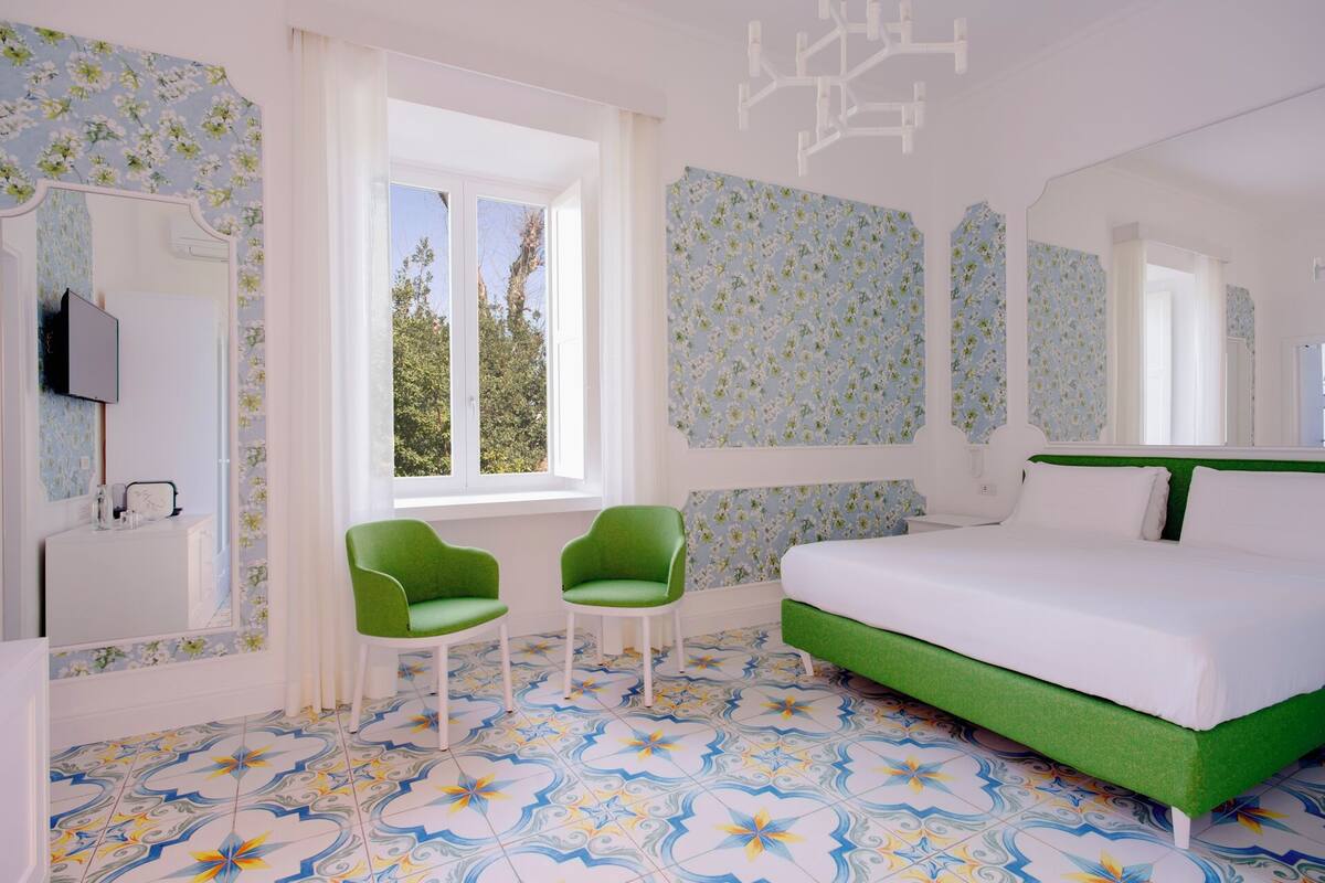 A bedroom with a bed, two green chairs, a TV, and a window with a view of trees.