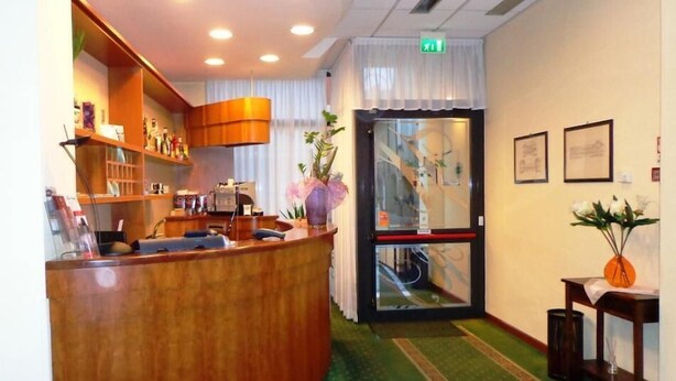 A hotel reception area with a wooden reception desk, a green carpet, and a glass door leading to a room. - Hotel Select (Montecchio Emilia)